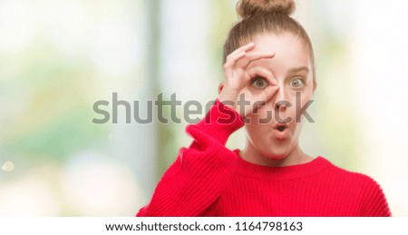 Young blonde woman wearing bun and red sweater doing ok gesture shocked with surprised face, eye looking through fingers. Unbelieving expression.