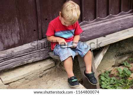 Cute 3-4 years old little boy sharing a photo on social media in internet sitting on the bench at old wooden house in village, summer outdoor, social network, gadget and technology concept