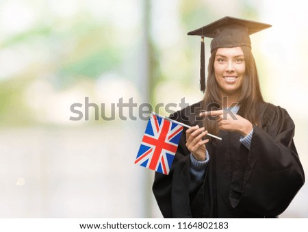Young hispanic woman wearing graduated uniform holding flag of united kingdom very happy pointing with hand and finger