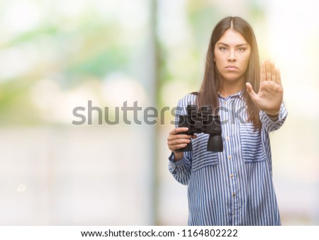 Young hispanic woman holding binoculars with open hand doing stop sign with serious and confident expression, defense gesture