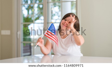 Down syndrome woman at home holding flag of usa with happy face smiling doing ok sign with hand on eye looking through fingers