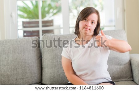 Down syndrome woman sitting on the sofa at home happy with big smile doing ok sign, thumb up with fingers, excellent sign