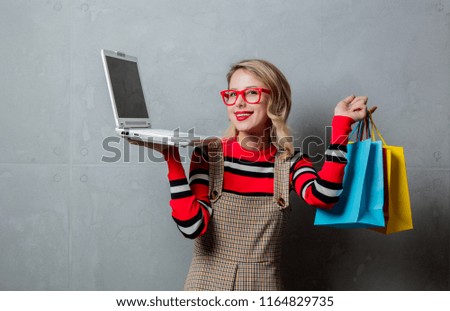 Portrait of a young style blonde girl in red striped sweater with shopping bags and laptop computer on grey background.