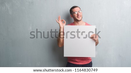 Young caucasian man over grey grunge wall holding a blank banner doing ok sign with fingers, excellent symbol