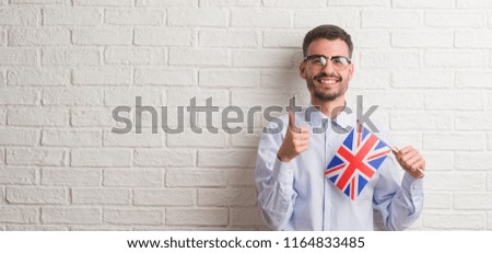 Young adult man over brick wall holding flag of United Kingdom happy with big smile doing ok sign, thumb up with fingers, excellent sign