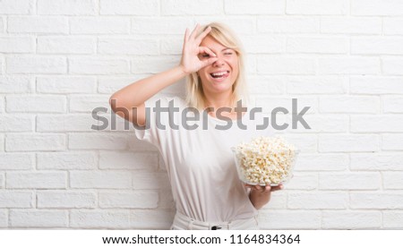 Adult caucasian woman over white brick wall eating pop corn with happy face smiling doing ok sign with hand on eye looking through fingers