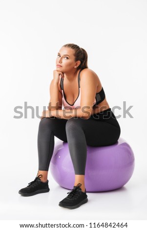 Beautiful plump girl in sporty top and leggings sitting on fitness ball thoughtfully looking aside over white background