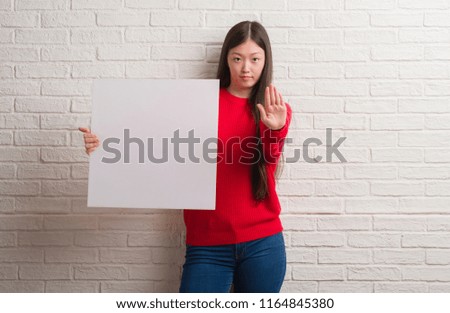 Young Chinese woman over brick wall holding banner with open hand doing stop sign with serious and confident expression, defense gesture