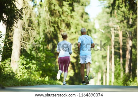 Back view of aged spouses in activewear running down road in park between trees on sunny day