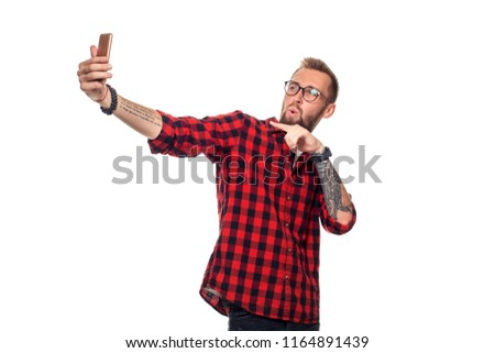 Lifestyle concept: a young man with a beard in shirt holding mobile phone and making photo of himself while standing against white background.