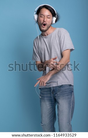 Young bearded man listening to music in blue background. Extremely excited Asian young hipster using white headphone to listen and dance to music, half body shot. Young generation hipster concept.