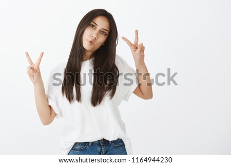 Chill and be positive. Portrait of joyful happy girl in casual outfit, raising hands in victory or peace gesture, sucking lips and making fish face, standing against gray background childish