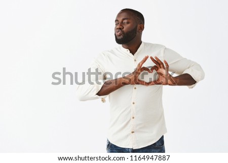 Come to me my love. Passionate and flirty funny dark-skinned guy with beard in casual outfit showing heart sign over chest folding lips in kiss and staring at camera, flirting over gray background