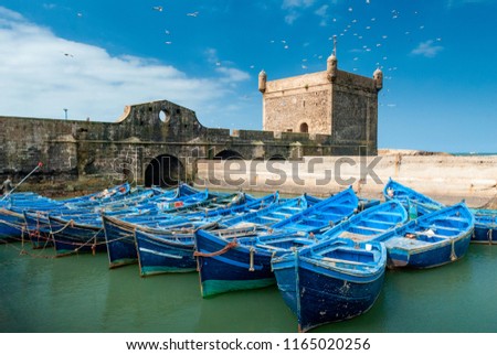 A fleet of blue fishing boats huddled together in the port of Essaouira in Morocco. You can also see the fortifications and a tower of the citadel of Mogador Royalty-Free Stock Photo #1165020256