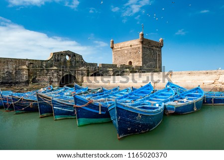 A fleet of blue fishing boats huddled together in the port of Essaouira in Morocco. You can also see the fortifications and a tower of the citadel of Mogador Royalty-Free Stock Photo #1165020370