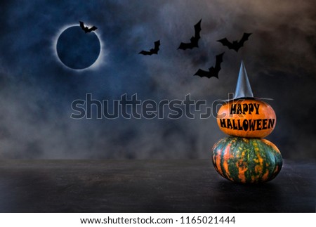 Two pumpkins with witch hat and text HAPPY HALLOWEEN against the background of the night sky, bats and the moon eclipse. Pumpkins At Moonlight In The Spooky Night - Halloween Scene Royalty-Free Stock Photo #1165021444