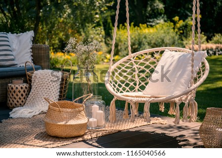 A beige string swing with a pillow on a patio. Wicker baskets, a rug and a blanket on a wooden deck in the garden. Royalty-Free Stock Photo #1165040566