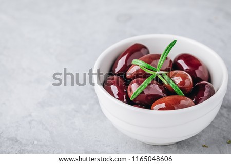 Fresh organic black olives in olive oil in white bowl with rosemary on gray stone background. 