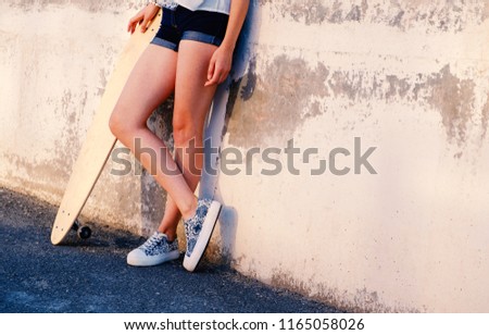 Bare legs of skater girl in short shorts leaning back used wall with her longboard near with her legs crossed a lot of space for text