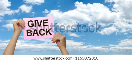 Make someone's day and GIVE BACK - female hands holding high a pink placard that says GIVE BACK against a wide blue sky and fluffy cloud summer background
