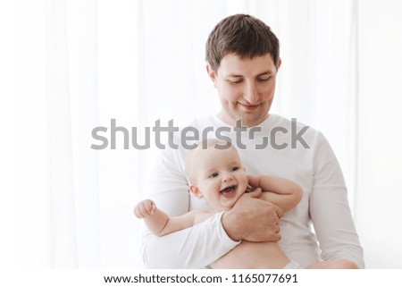 Handsome man smiling and holding funny baby while standing on background of white curtains