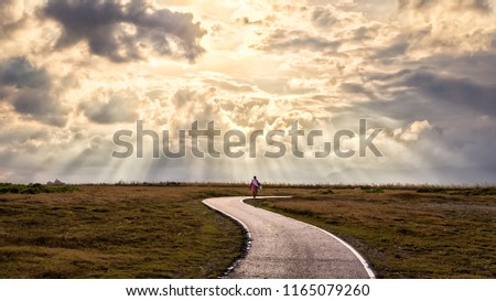 A person walking alone along s-shape path. The sun produces amazing light rays across the sky. The image is simple and breathtaking. This image is suitable for background use or add quote above. Royalty-Free Stock Photo #1165079260