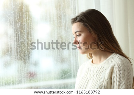 Sad woman looking outdoors through a window in a rainy day at home Royalty-Free Stock Photo #1165183792