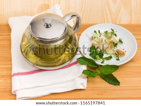 Herbal tea in glass teapot with strainer on napkin and some herbs on saucer and beside on a wooden surface
