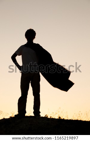 Boy playing superheroes on the sky background, teenage superhero in a red cloak on a hill