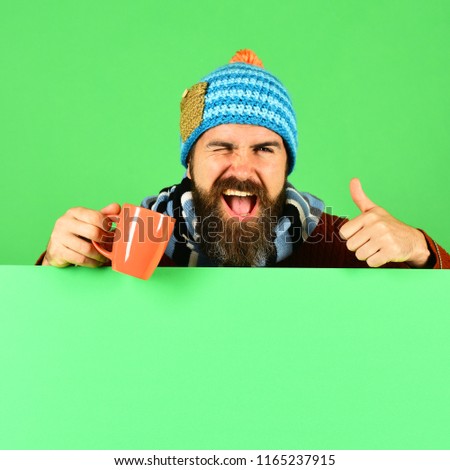 Man in hat holds brown cup on green background, copy space. October beverage idea. Autumn and hot drink season concept. Hipster with beard and flirty face holds tea or coffee cup and shows thumbs up