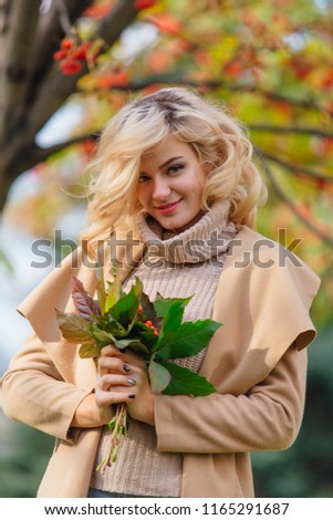 Beautiful elegant blonde woman dressed in a coat standing under the rowan tree holding bouquet of rowanberries and leaves in autumn park. Sunny october day.