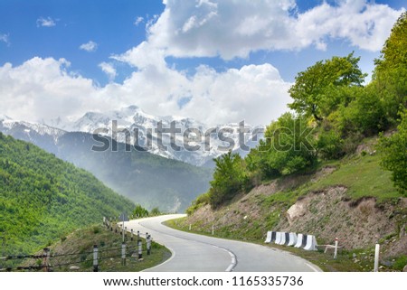 Empty road serpentine in the mountains, blue cloudy sky, mountain peaks in the snow and green hills background