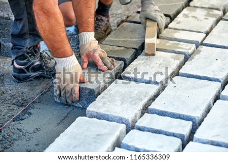 A workman's gloved hands use a hammer to place stone pavers. Worker creating pavement using cobblestone blocks and granite stones.