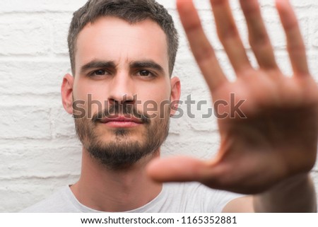 Young adult man standing over white brick wall with open hand doing stop sign with serious and confident expression, defense gesture