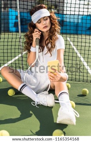 stylish sportswoman in white sportswear with tennis racket taking selfie on smartphone at net on tennis court