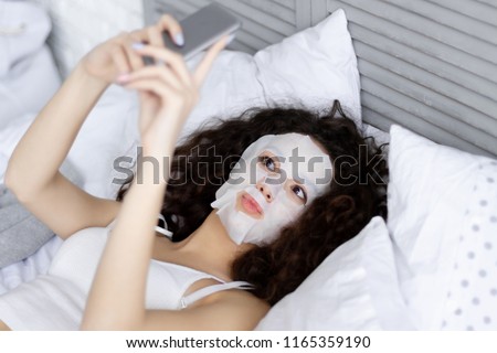 Brunette cute young girl female woman lying in bed with refreshing spa body care mask doing selfie in light room at home. Morning wake up / body care concept