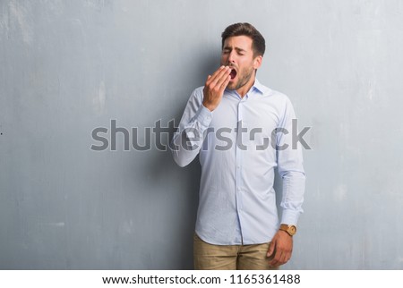 Handsome young business man over grey grunge wall wearing elegant shirt bored yawning tired covering mouth with hand. Restless and sleepiness.