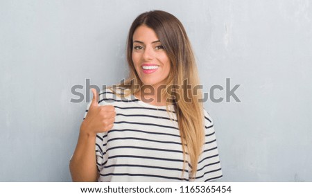 Young adult woman over grunge grey wall wearing navy t-shirt doing happy thumbs up gesture with hand. Approving expression looking at the camera with showing success.