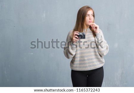 Young adult woman over grey grunge wall taking pictures using vintage camera serious face thinking about question, very confused idea