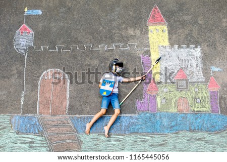 Little active kid boy drawing knight castle and fortress with colorful chalks on asphalt. Happy child in helmet and with spear having fun with playing knight game and painting