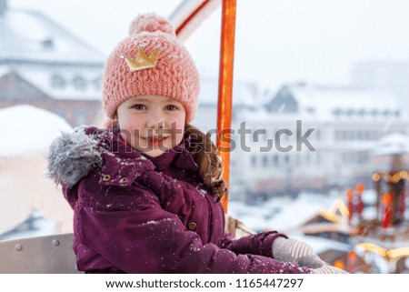 Little cute kid girl having fun on ferris wheel on traditional German Christmas market during strong snowfall. Happy child enjoying family market in Germany, Munich. Laughing girl on carousel: