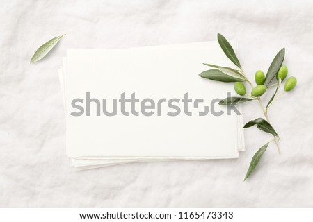 minimalist feminine styled invitation card mock-up (hand made paper) with fresh olive twig on a soft white linen background - perfect for wedding stationery. Flat lay / top view.