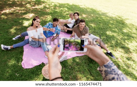 friendship, leisure and technology concept - group of happy smiling friends with non alcoholic drinks taking picture by smartphone on picnic at summer park