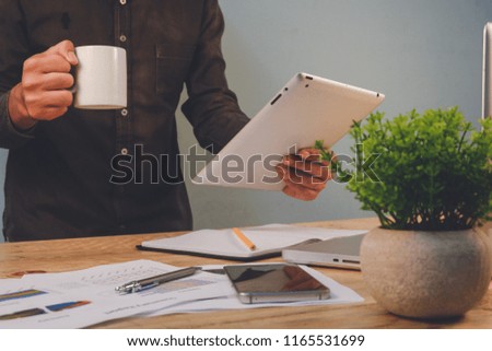 Young business man sitting in office at wooden table and using tablet . On desk is laptop and smartphone on hand holding coffee,  man analyzing data report . Student learning online.