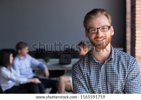 Portrait of smiling red haired male employee look at camera during business meeting in office, happy man in glasses pose for corporate photoshoot, worker or team leader making picture during briefing