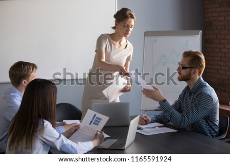 Millennial businesswoman give handout materials to work team members during flipchart presentation, female speaker or coach share documents to workers, presenter hand papers to consider or analyze Royalty-Free Stock Photo #1165591924