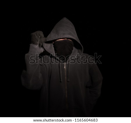 Hooded Masked Thug with a Knife Raised in a Stabbing Motion. Dark Moody Black Background. Knife crime inner city gangs. Assault and robbery. An assassin, murderer or protester in violent assault. Royalty-Free Stock Photo #1165604683