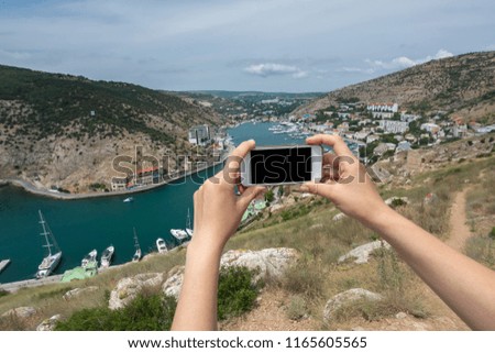 Horizontal smartphone in the hand with blank screen. On the background small resort town with small bay full of ships in blue sea water.