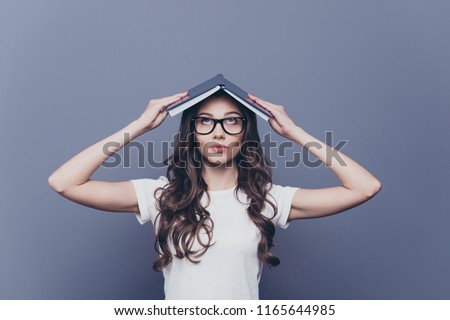 Attractive smart, clever, lovely stylish nice cute curly-haired brunette girl in casual white t-shirt and glasses, holding opened book on head like house, isolated on grey background