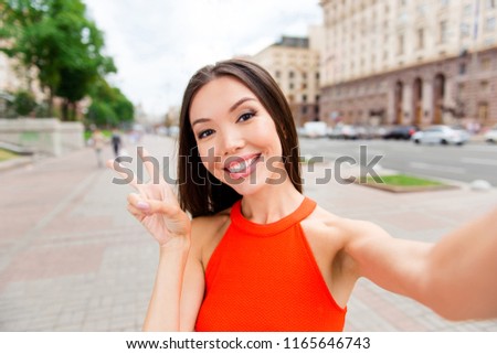Pretty young woman making selfie on smartphone and showing two fingers like v-sign look into the camera